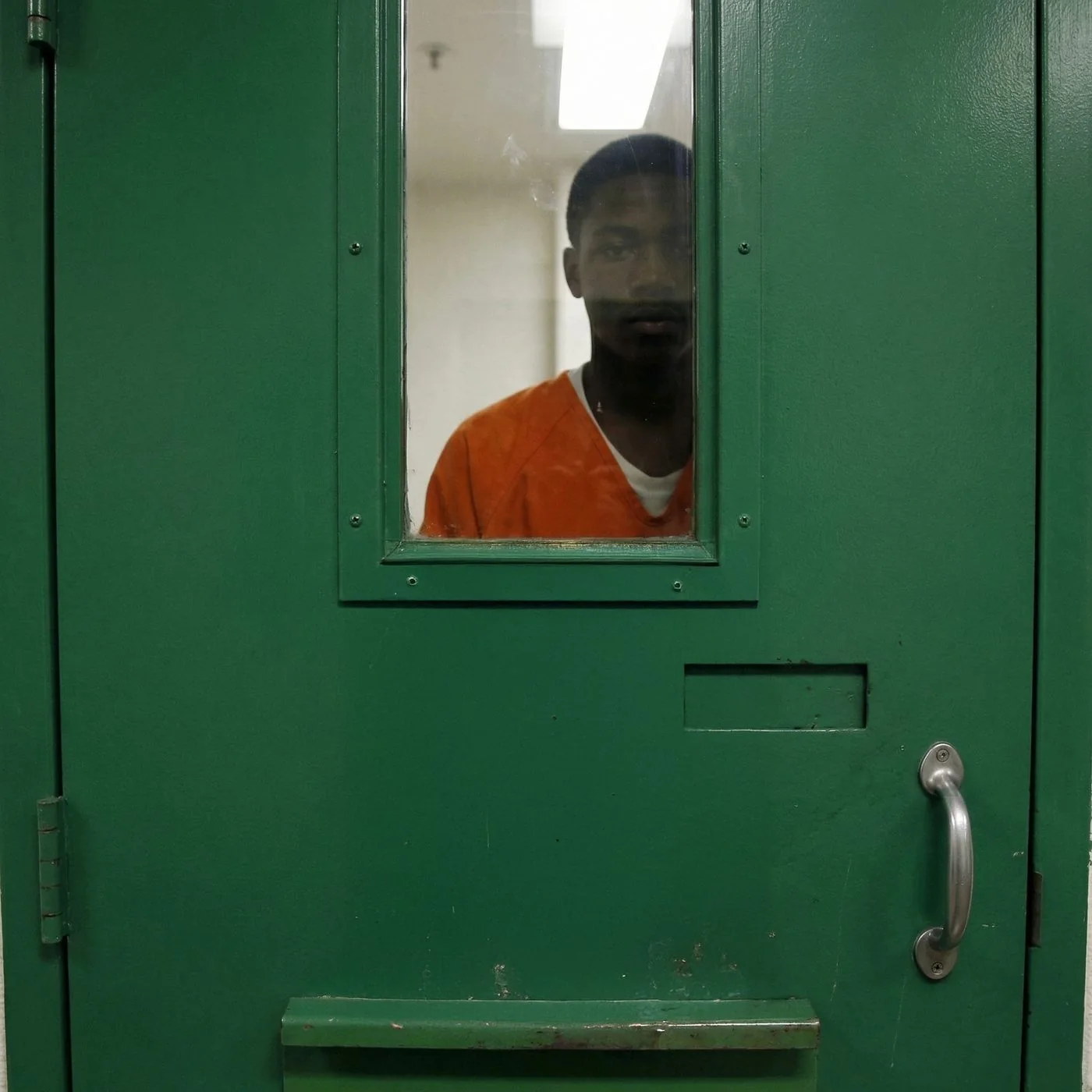A young man in an orange jumpsuit looks through the window of a green cell door