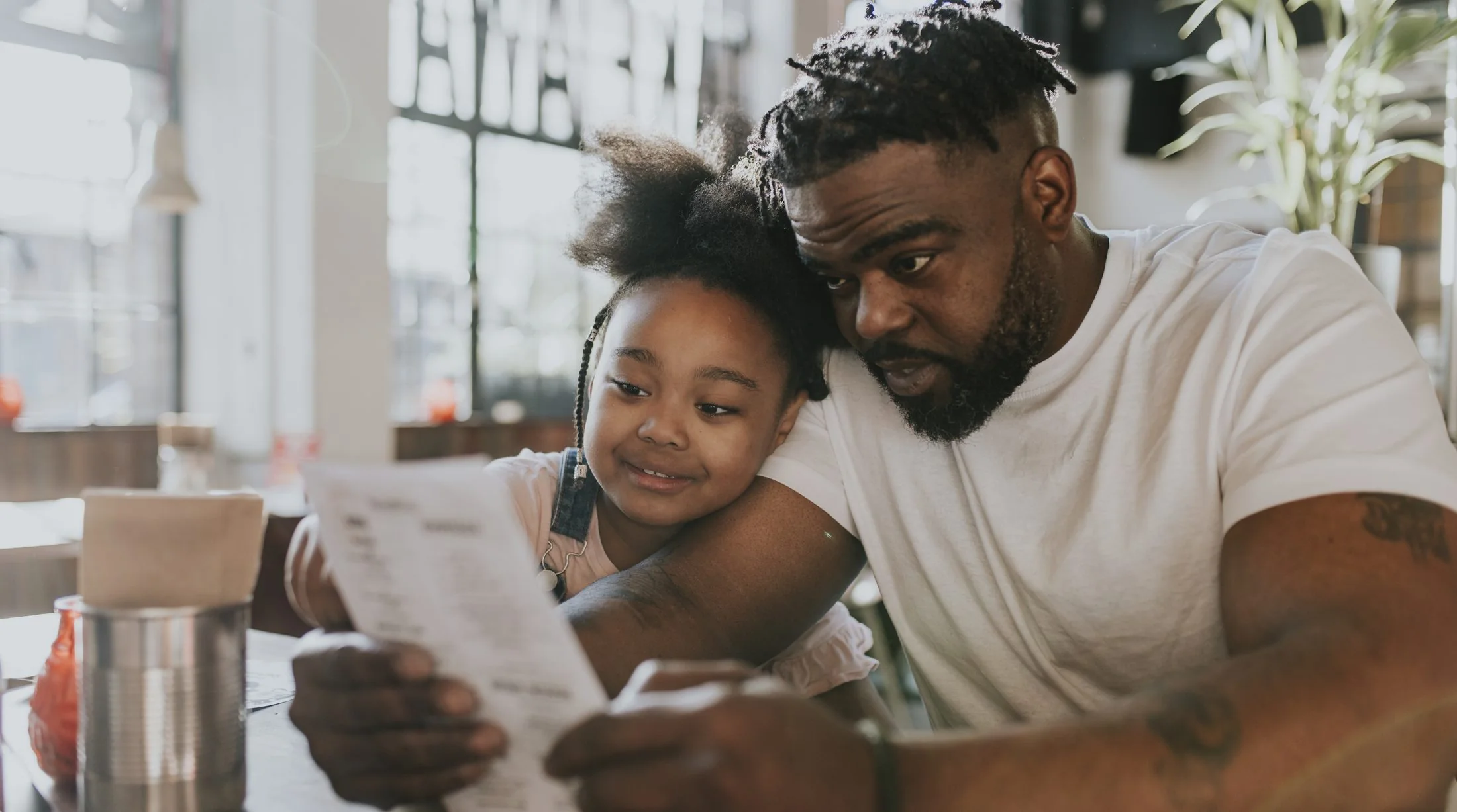 A father reads with his young daughter at a cafe table