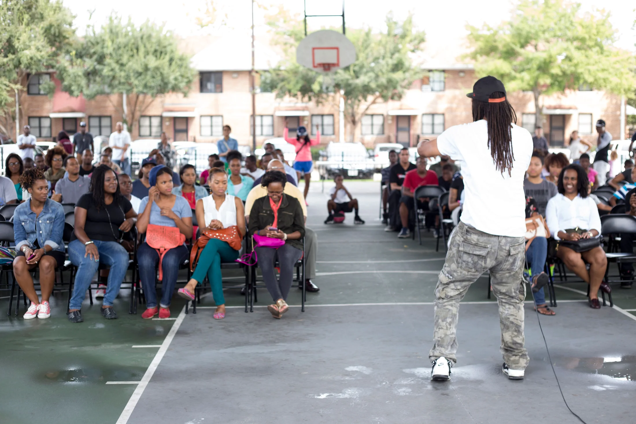 A crowd seated in folding chairs on an outdoor basketball court as Ronnie Lillard speaks to them