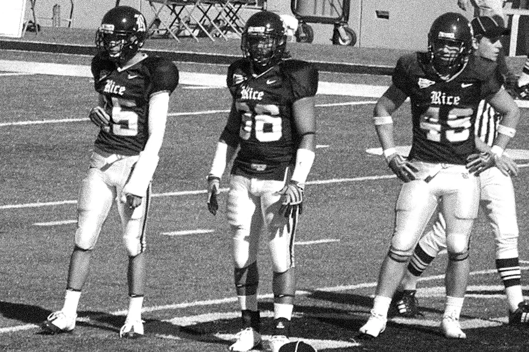 Three Rice football players in dark jerseys on the sideline, Ronnie Lillard among them