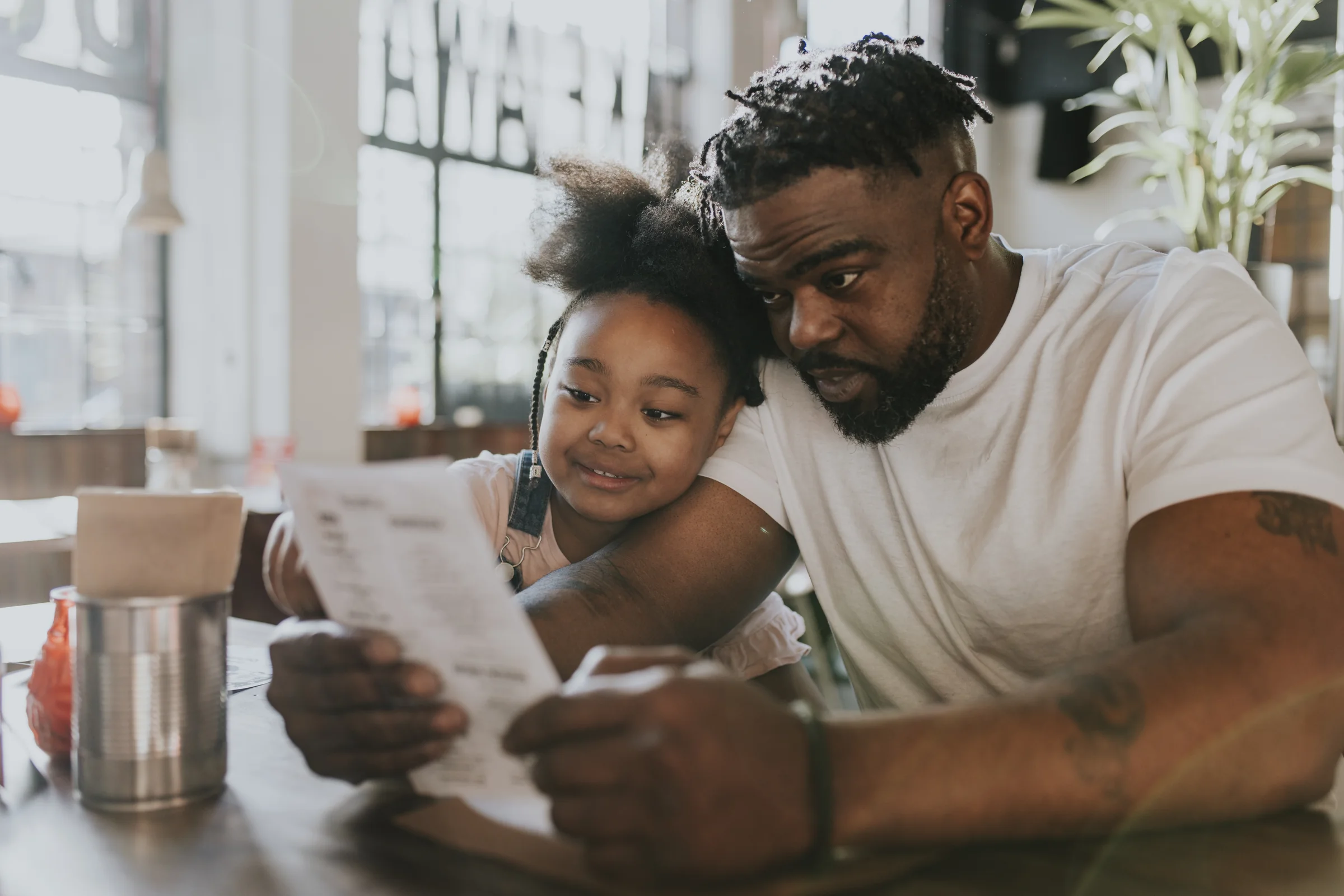A father and his young daughter read a menu together at a cafe table, her head resting on his arm