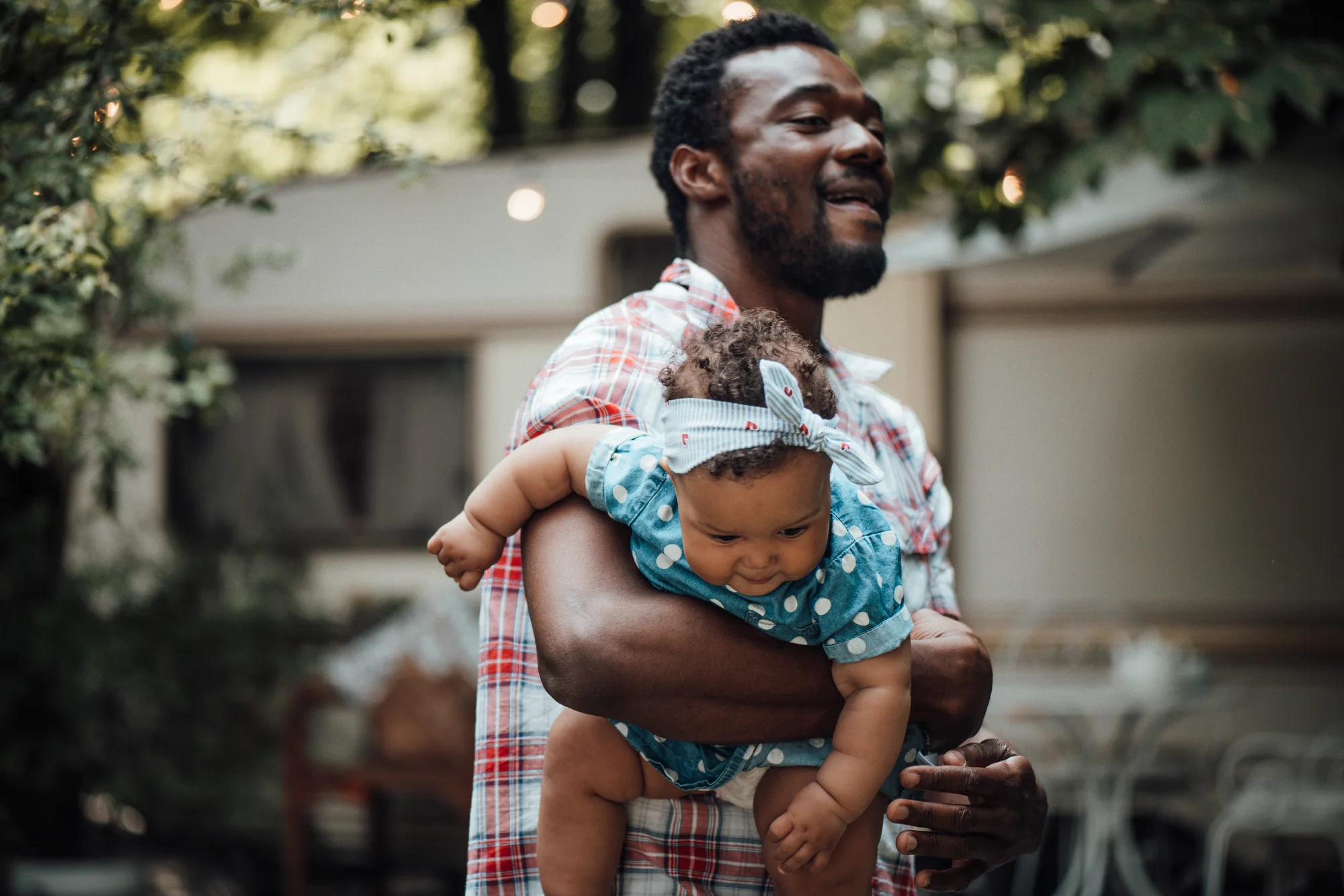 A laughing father carries his baby daughter in a polka dot outfit and headband under string lights outdoors