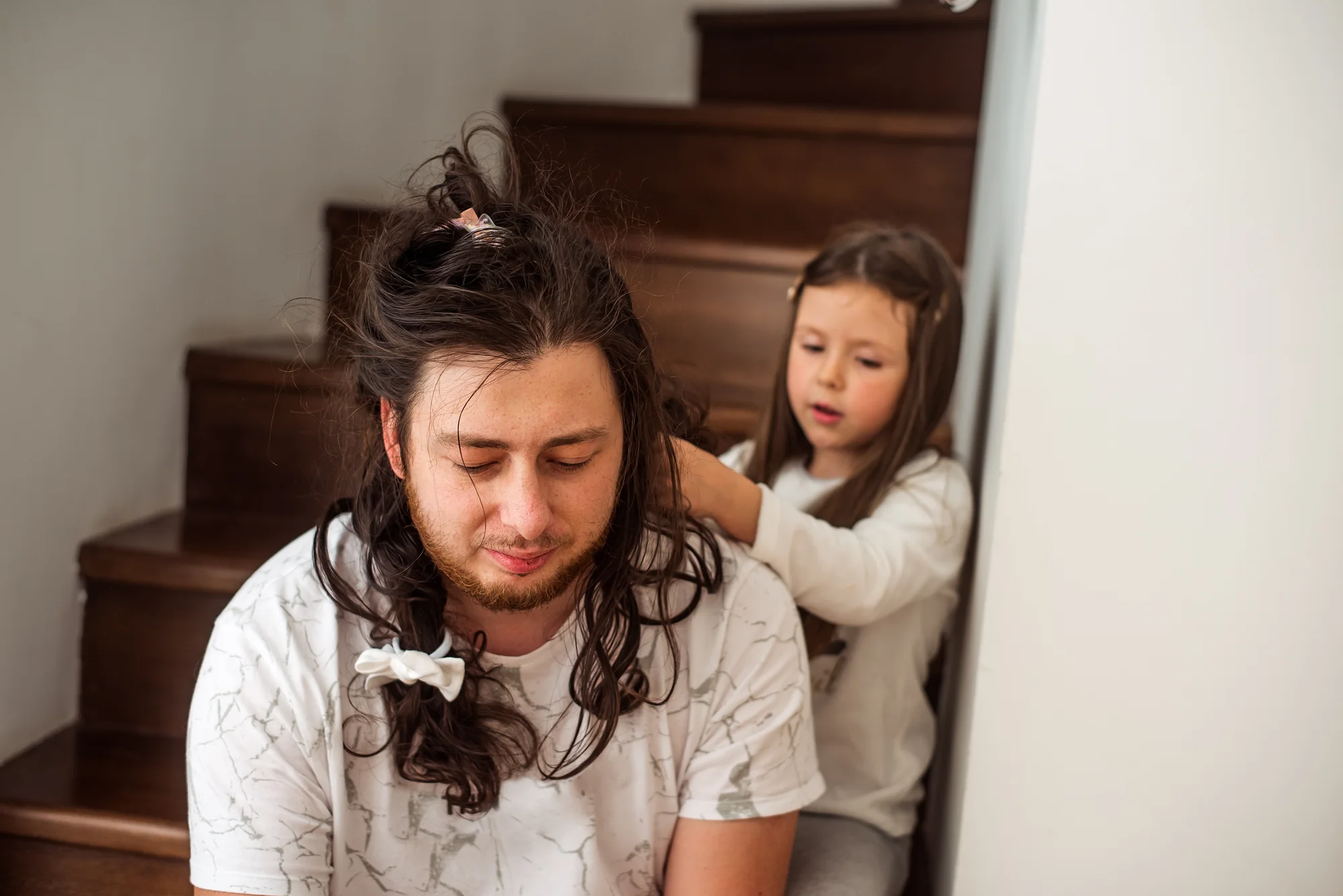 A young girl styles her father’s long hair with clips and a bow as he sits on the stairs, eyes closed